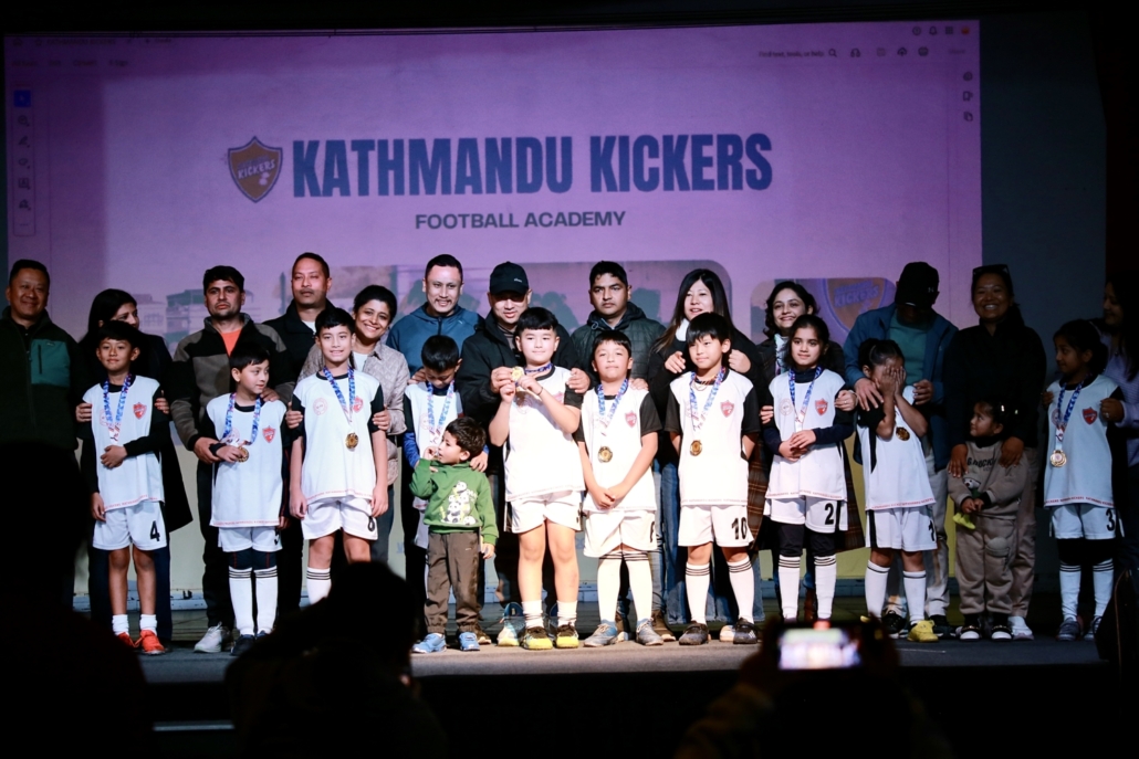 Group photo of young football players and coaches from Kathmandu Kickers Football Academy, standing on stage with medals, celebrating together with parents and supporters.